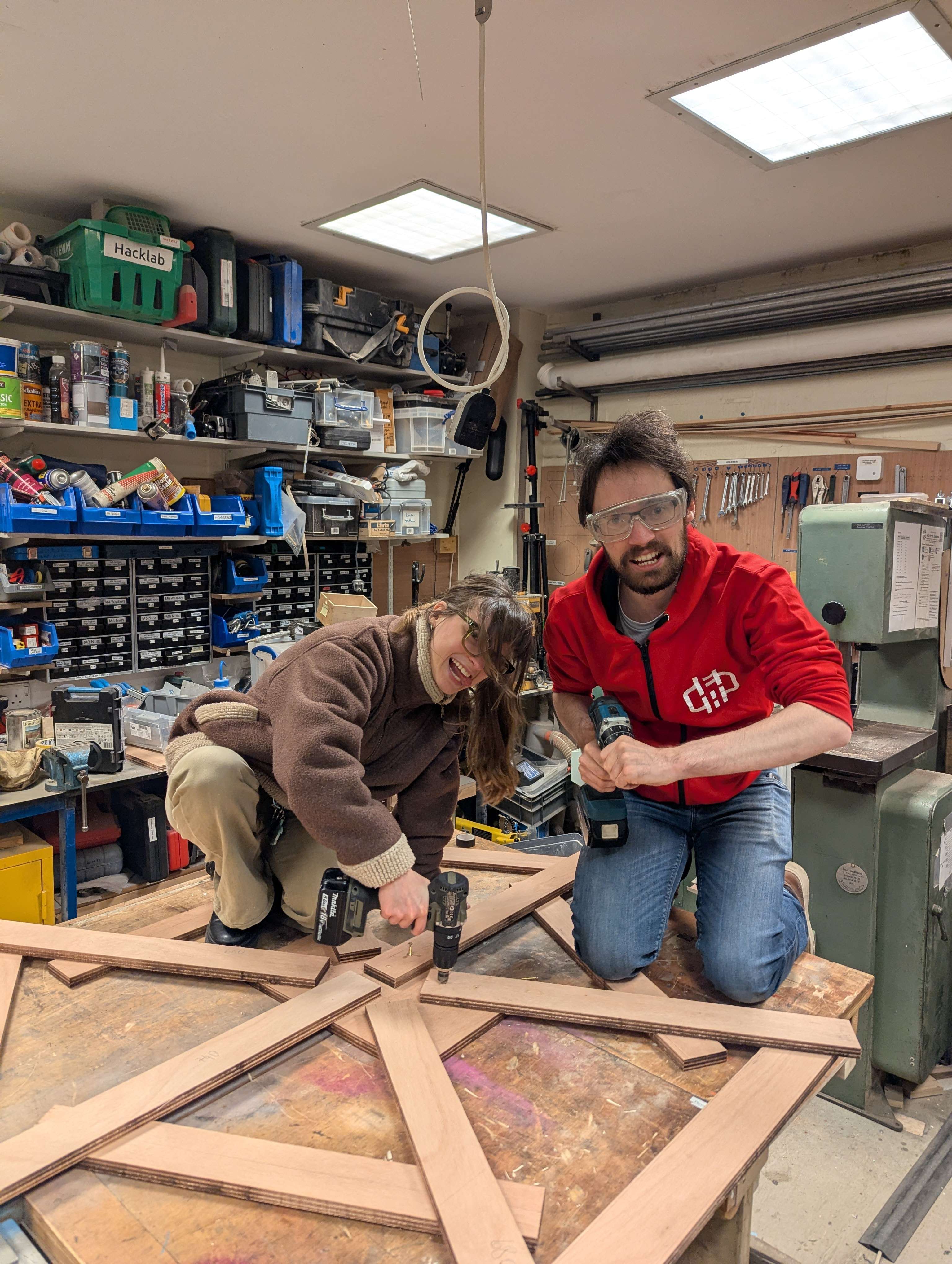 Two people kneeling on top of a workbench with a wooden structure on it. One is drilling into the structure.