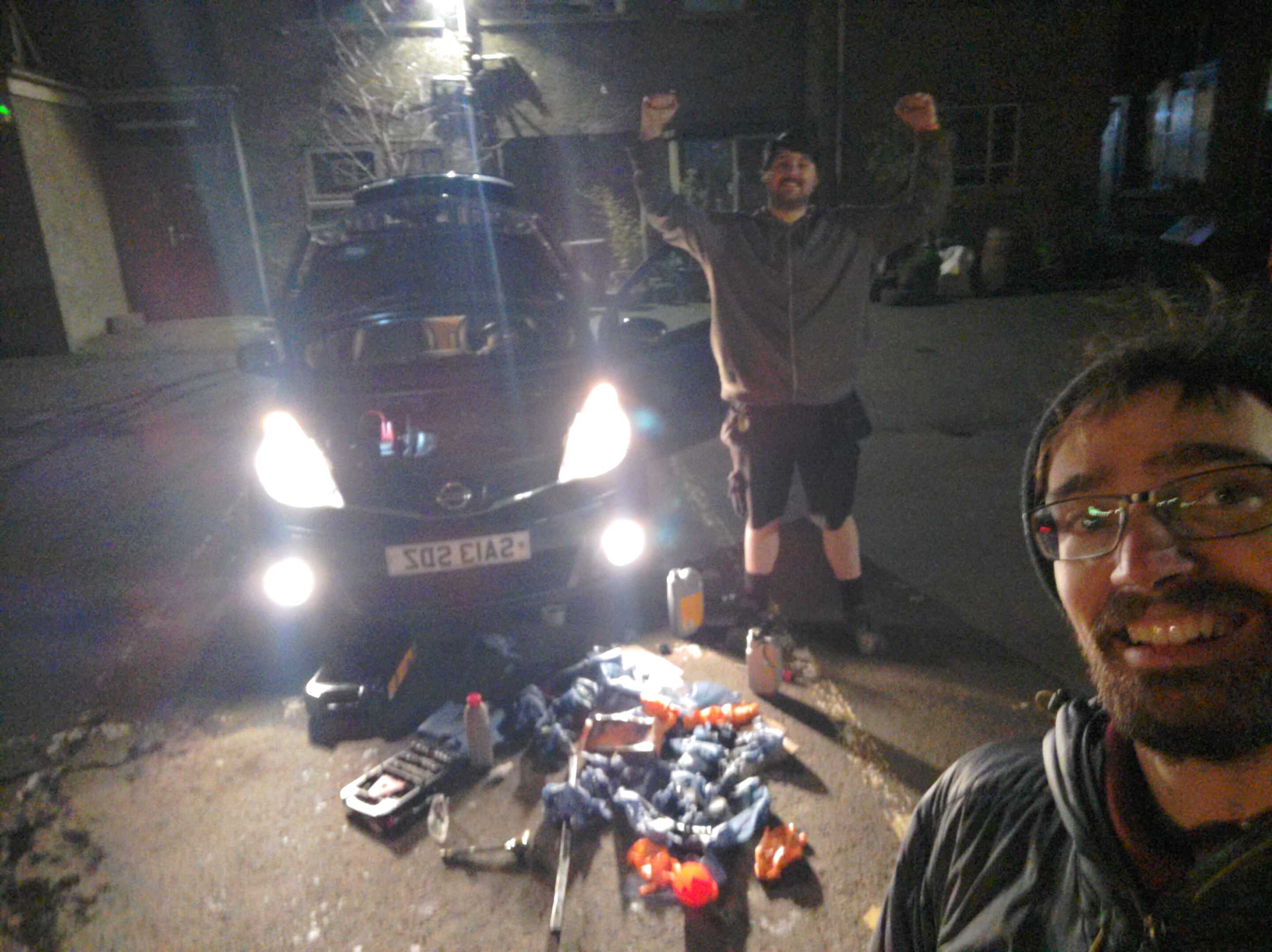 Two people at the camera in front of a car. Tools are scattered in front of the car, and one person is cheering.